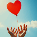 Close-up of gentle hands releasing a red heart-shaped balloon into a bright blue sky, symbolizing letting go and healing, representing the emotional journey in how to get over someone.