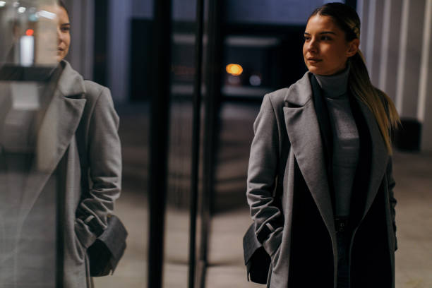 A stylish lady looking at her reflection in an outdoor mirror at night, showcasing how to dress for a date with a polished monochrome outfit and elegant accessories.